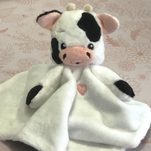 以下が含まれることがあります： ピンクのハートがついた白黒の牛のぬいぐるみ。ぬいぐるみはベビー用のおくるみです。