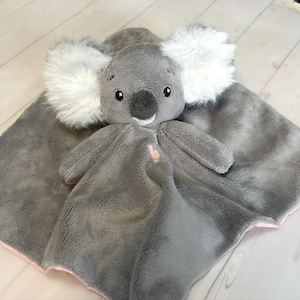 May include: A gray plush koala bear with white fluffy ears and a smiling face, attached to a gray blanket with a pink trim. The koala has a small pink heart on its chest. Soft toy for babies.