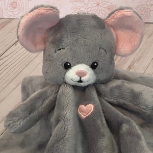 Könnte beinhalten: Eine graue Plüschmaus-Sicherheitsdecke mit rosa Ohren und einem rosa Herz auf der Vorderseite.