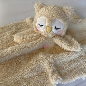May include: A cream-coloured baby security blanket with an owl face, closed eyes, pink cheeks, and a yellow beak. The blanket has a soft, textured surface and a small pink heart accent.