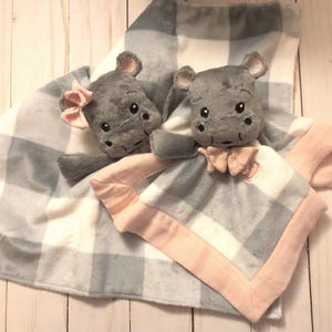 Könnte beinhalten: Eine graue und weiße karierte Babydecke mit rosa Rand. Zwei graue Plüsch-Nilpferd-Spielzeuge sind an der Decke befestigt.