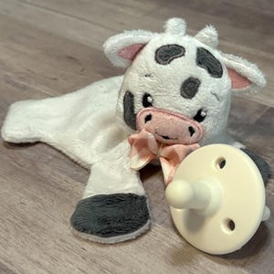 Op de afbeelding: Een witte, pluche koe-vormige babyknuffel met zwarte vlekken, grijze hoeven en een roze ruitjesstrik. Een crèmekleurige fopspeen ligt naast de knuffel. De koe heeft een roze neus en oren.