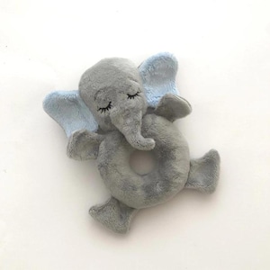以下が含まれることがあります： 目を閉じたグレーとブルーのぬいぐるみ象のリングおもちゃ。象は柔らかくふわふわの生地で作られており、赤ちゃんが持ったり遊んだりできる中央にリングがあります。