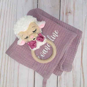May include: A soft, mauve muslin baby blanket with a white lamb plush toy attached to a wooden teething ring. The blanket is embroidered with the name "Caroline".