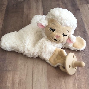 May include: White plush lamb with closed eyes and a pink nose. The lamb is lying down with its head resting on its paw. A pacifier is attached to the lamb's paw.