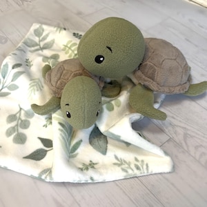 Op de afbeelding: Twee pluche schildpadden met zachte olijfgroene koppen en ledematen, voorzien van lichtbruine, getextureerde schilden. De schildpadden rusten op een witte stoffen deken met een herhalend patroon van saliegroene bladeren en botanische motieven.