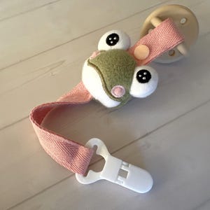 May include: A baby pacifier clip featuring a plush frog head in green, white, and pink. The clip has a pink fabric strap and a white plastic clip. The pacifier is light brown.