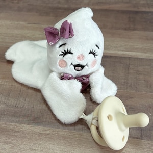 May include: A white plush ghost-shaped baby toy with embroidered facial features, a purple bow, and a matching pacifier. The ghost has a smiling expression and is lying down. The pacifier is attached with a white cord.