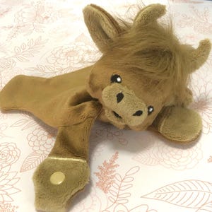 May include: A brown plush toy in the shape of a Highland cow with a soft, fuzzy texture. The toy has a stitched face with black eyes and a black nose. The toy has two small, brown, fabric loops on the back of its body.