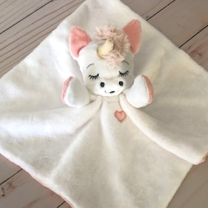 Può includere: Coperta di sicurezza in peluche bianca a forma di unicorno con dettagli rosa e un cuore rosa ricamato sul davanti.