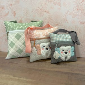 May include: Three quilted fabric pouches in varying sizes and colours. The largest is light green with a diamond pattern, the medium is pink with a floral pattern, and the smallest is grey with a gingham pattern. Each pouch features a zip closure and a smiling tooth applique.