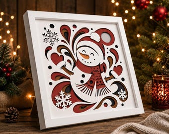 May include: A square white frame containing a multi-layered paper cut art piece. The design features a central snowman wearing a red hat and scarf with mittens, surrounded by white snowflakes and swirling red, black, and tan abstract shapes.
