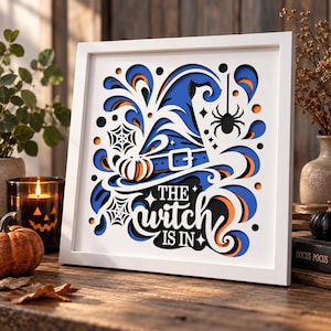 Puede incluir: Una pieza de arte en papel superpuesto con marco blanco cuadrado que presenta un sombrero de bruja azul, una araña negra, telarañas y detalles en naranja. El texto central dice THE witch IS IN en letras blancas sobre un fondo oscuro.