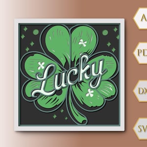 Puede incluir: Una pieza de arte en papel en capas con un trébol verde con la palabra "Lucky" en una fuente de escritura. El trébol está sobre un fondo gris oscuro con detalles en blanco, enmarcado en un cuadrado blanco.
