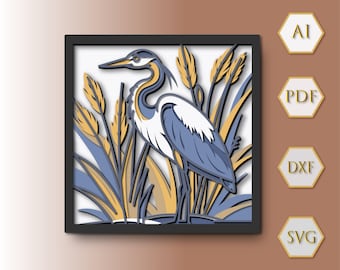 May include: A layered paper art piece featuring a blue heron standing in tall grass. The artwork is framed in a black square frame. The heron has white, blue, and gray feathers. The grass is yellow and blue. The background is white.