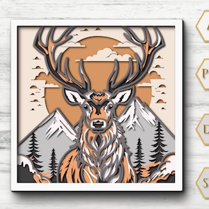 May include: A layered paper art piece featuring a majestic stag with large antlers, set against a backdrop of mountains, trees, and a setting sun. The artwork uses shades of brown, gray, and white, creating a 3D effect within a white square frame.
