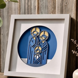 Puede incluir: Un marco cuadrado blanco que contiene una obra de arte de papel en capas. La pieza presenta una representación estilizada de tres figuras en tonos azules y amarillos, sobre un fondo circular azul oscuro, creando un efecto dimensional.