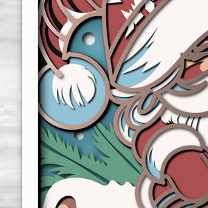 May include: A multi-layered paper art piece featuring a stylized Santa Claus figure with a white beard, red hat, and red clothing. The design incorporates light blue, dark teal, and green pine branch elements, creating depth through stacked cutouts.