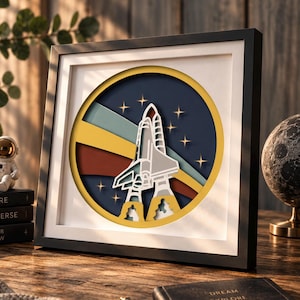 Op de afbeelding: Een vierkante zwarte lijst met een meerlaags papieren kunstwerk. Het cirkelvormige ontwerp toont een witte spaceshuttle die opstijgt tegen een donkerblauwe achtergrond met gele sterren. Achter de shuttle bevinden zich horizontale strepen in geel, rood en blauwgroen.