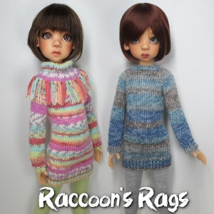 May include: Two dolls wearing knitted sweaters. The doll on the left is wearing a pink, yellow, and blue striped sweater with a fringe trim. The doll on the right is wearing a blue and gray striped sweater.  Raccoon's Rags.