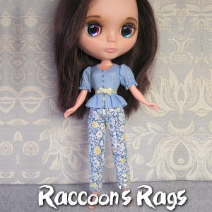 May include: A doll wearing a light blue blouse with a yellow ribbon and matching blue and white floral pants. The doll has long dark hair and blue eyes with purple and green accents. The doll is standing on a light blue and white patterned background. The text "Raccoons Rags" is at the bottom of the image.