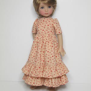 K&ouml;nnte beinhalten: Puppe mit blonden Haaren und blauen Augen, bekleidet mit einem cremefarbenen, langen Kleid mit rotem Blumenmuster. Das Kleid hat kurze &Auml;rmel und gestufte R&uuml;schen. Die Puppe steht vor einem wei&szlig;en Hintergrund.