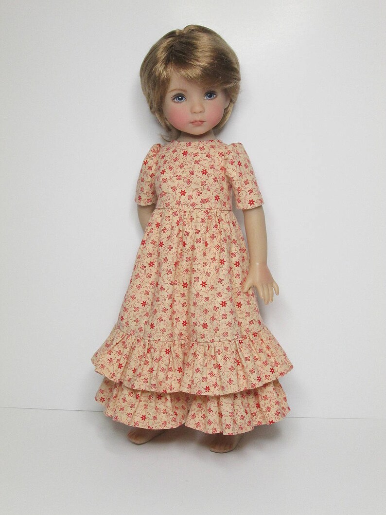 K&ouml;nnte beinhalten: Puppe mit blonden Haaren und blauen Augen, bekleidet mit einem cremefarbenen, langen Kleid mit rotem Blumenmuster. Das Kleid hat kurze &Auml;rmel und gestufte R&uuml;schen. Die Puppe steht vor einem wei&szlig;en Hintergrund.