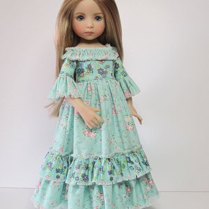 K&ouml;nnte beinhalten: Eine Puppe mit langen blonden Haaren, die ein hellblaues Kleid mit Blumenmuster und R&uuml;schenbesatz tr&auml;gt.