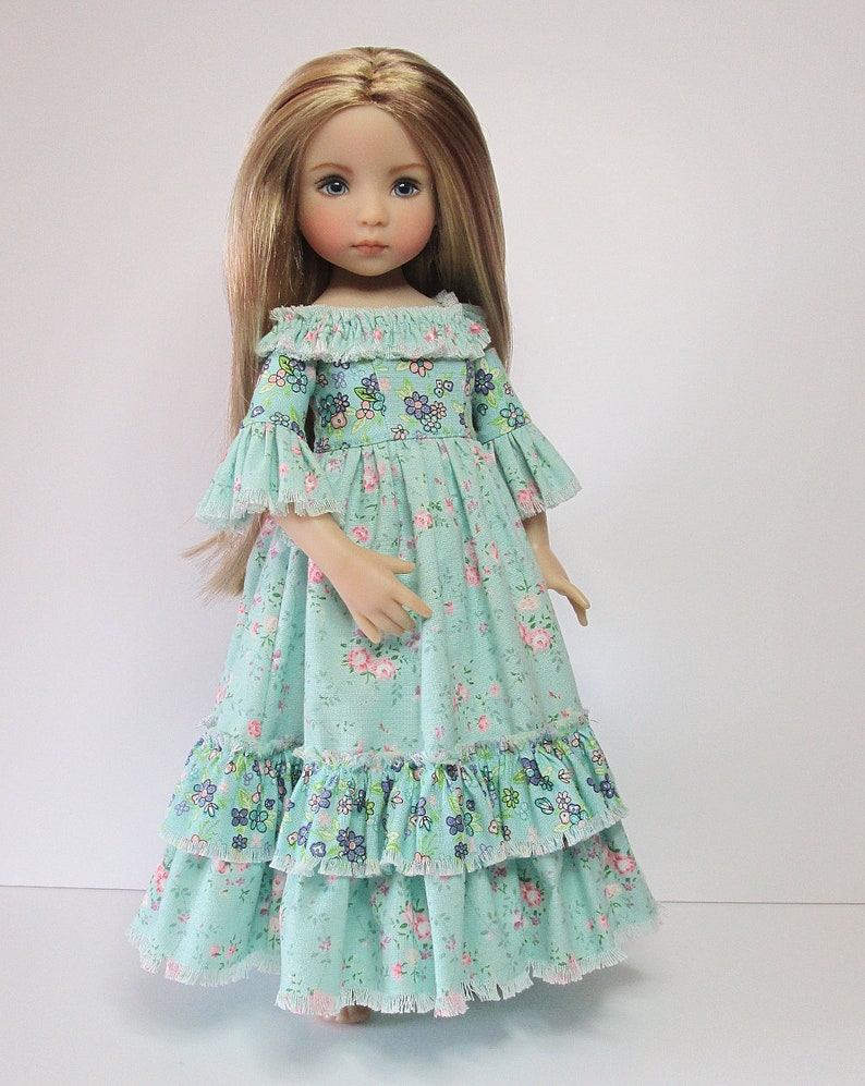 K&ouml;nnte beinhalten: Eine Puppe mit langen blonden Haaren, die ein hellblaues Kleid mit Blumenmuster und R&uuml;schenbesatz tr&auml;gt.