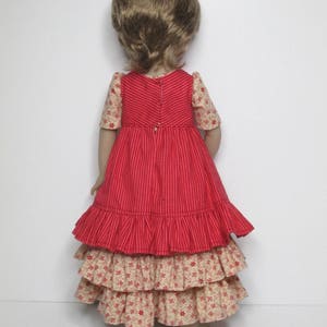K&ouml;nnte beinhalten: Eine Puppe in einem rot-cremefarbenen Kleid. Das Oberteil ist rot mit vertikalen Streifen, und die &Auml;rmel sind cremefarben mit einem Blumenmuster. Der Rock hat gestufte R&uuml;schen in Creme mit einem roten Blumendruck. Die Puppe hat hellbraunes Haar.