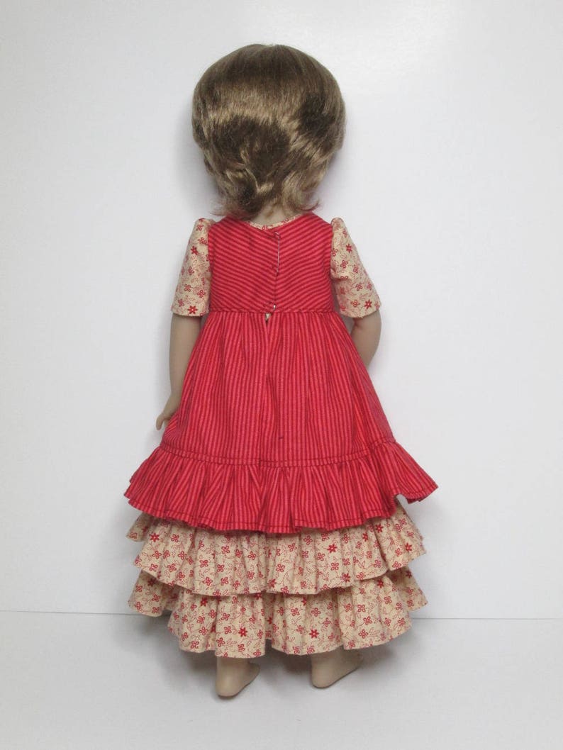 K&ouml;nnte beinhalten: Eine Puppe in einem rot-cremefarbenen Kleid. Das Oberteil ist rot mit vertikalen Streifen, und die &Auml;rmel sind cremefarben mit einem Blumenmuster. Der Rock hat gestufte R&uuml;schen in Creme mit einem roten Blumendruck. Die Puppe hat hellbraunes Haar.