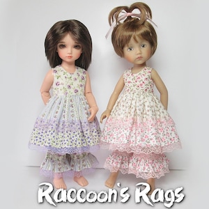 Puede incluir: Dos muñecas con vestidos de estampado floral con ribetes de encaje. La muñeca de la izquierda lleva un vestido lavanda y blanco con una falda a juego. La muñeca de la derecha lleva un vestido rosa y blanco con una falda a juego. Ambas muñecas tienen el pelo castaño y los ojos azules. Las muñecas están de pie sobre un fondo blanco. Raccoon's Rags.