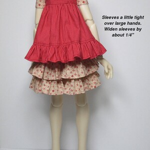 K&ouml;nnte beinhalten: Ein rotes und cremefarbenes Puppenkleid mit gestreiftem Oberteil und ger&uuml;schtem Rock. Die &Auml;rmel sind mit kleinen roten Blumen gemustert. Der Text auf dem Bild lautet "Fits Kaye Wiggs Tobi body."