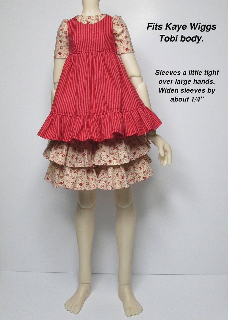 K&ouml;nnte beinhalten: Ein rotes und cremefarbenes Puppenkleid mit gestreiftem Oberteil und ger&uuml;schtem Rock. Die &Auml;rmel sind mit kleinen roten Blumen gemustert. Der Text auf dem Bild lautet "Fits Kaye Wiggs Tobi body."