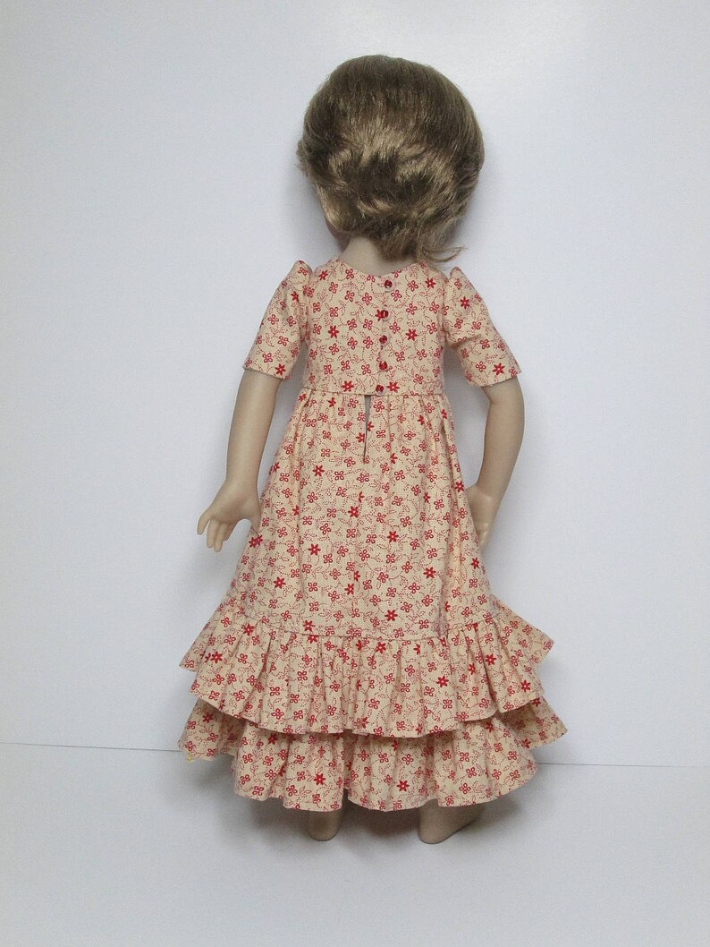 K&ouml;nnte beinhalten: Puppe in einem cremefarbenen Kleid mit rotem Blumenmuster. Das Kleid hat kurze &Auml;rmel, ein figurbetontes Oberteil und einen gestuften, ger&uuml;schten Rock. Die Puppe hat kurze, hellbraune Haare.