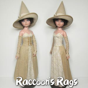 Op de afbeelding: Twee poppen die beige en witte jurken dragen met gouden accenten. Beide poppen dragen grote, puntige, beige hoeden met gouden accenten. De poppen staan op een witte achtergrond. De tekst "Raccoon's Rags" is onderaan de afbeelding gedrukt.