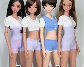 May include: Four dolls wearing different outfits. Each doll is wearing a white short-sleeved shirt with a different coloured bottom. The doll on the left is wearing a lavender bottom with white lace trim. The doll in the middle is wearing a light blue bottom with white lace trim. The doll on the right is wearing a lavender bottom with white lace trim. The doll in the back is wearing a light blue bottom with white lace trim. The dolls are all standing in a row. The text "Raccoon's Rags" is printed at the bottom of the image.