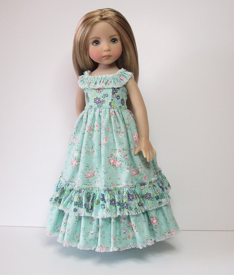 K&ouml;nnte beinhalten: Eine Puppe tr&auml;gt ein hellblaues Kleid mit Blumenmuster und mehreren R&uuml;schen. Das Kleid hat einen wei&szlig;en Spitzenbesatz.