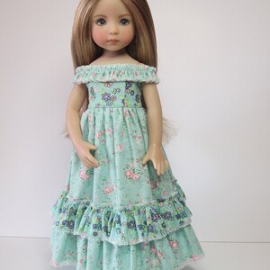 K&ouml;nnte beinhalten: Eine Puppe tr&auml;gt ein hellblaues Kleid mit Blumenmuster und mehreren R&uuml;schen. Das Kleid hat einen Ausschnitt mit offener Schulter.