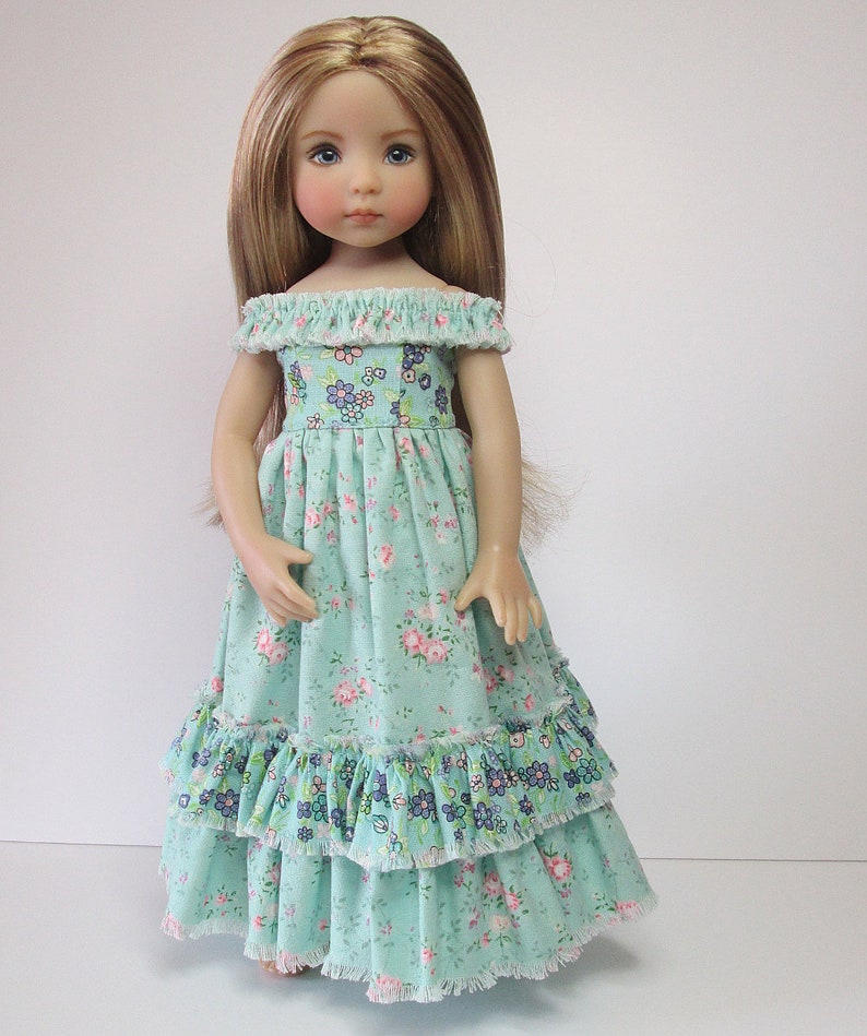 K&ouml;nnte beinhalten: Eine Puppe tr&auml;gt ein hellblaues Kleid mit Blumenmuster und mehreren R&uuml;schen. Das Kleid hat einen Ausschnitt mit offener Schulter.