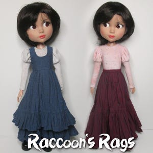 May include: Two dolls wearing different outfits. The doll on the left is wearing a blue dress with a white blouse. The doll on the right is wearing a burgundy dress with a pink blouse. Both dolls have brown hair and brown eyes. The text "Raccoon's Rags" is at the bottom of the image.