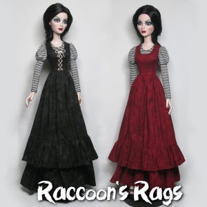 May include: Two dolls wearing long, tiered dresses with ruffled hems. The doll on the left is wearing a black dress with a white and black striped long-sleeved shirt. The doll on the right is wearing a red dress with a white and black striped long-sleeved shirt. Both dolls have black hair and are wearing a similar hairstyle. The text "Raccoon's Rags" is at the bottom of the image.