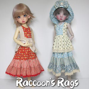May include: Two dolls wearing vintage-style dresses. The doll on the left is wearing a red, white and blue dress with a floral print. The doll on the right is wearing a yellow, blue and white dress with a floral print. Both dolls are wearing hats.