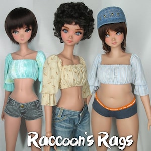 May include: Three dolls wearing different crop tops and denim shorts. The doll on the left is wearing a light blue crop top with a tie-dye pattern. The doll in the middle is wearing a yellow crop top with a floral pattern. The doll on the right is wearing a blue and white striped crop top.
