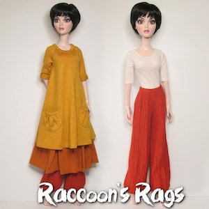 May include: Two dolls wearing different outfits. The doll on the left is wearing a long, mustard yellow tunic over a rust-colored skirt and red pants. The doll on the right is wearing a white, short-sleeved shirt and red, wide-legged pants. Both dolls have black hair and are wearing makeup. The text "Raccoon's Rags" is printed at the bottom of the image.