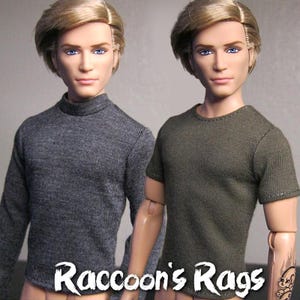 May include: Two dolls wearing different shirts. One doll is wearing a grey long-sleeved shirt and the other doll is wearing a green short-sleeved shirt. The text "Raccoon's Rags" is at the bottom of the image.