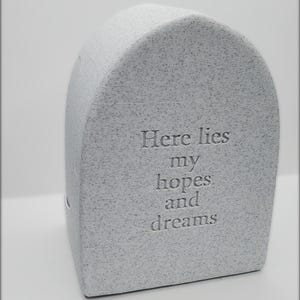 May include: A gray, speckled, stone-like decorative object with rounded top and flat base. The text "Here lies my hopes and dreams" is engraved on the front. The object has a simple, minimalist design.