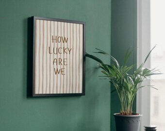 May include: A framed art print with the text "HOW LUCKY ARE WE" in a minimalist style. The artwork features vertical beige stripes on a cream background, encased in a black frame. The print hangs on a dark green wall.