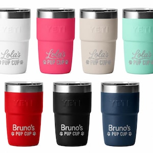 May include: Seven stainless steel Yeti tumblers in white, pink, beige, mint, red, black, and navy blue. Each features a metallic rim and is engraved with either Lola's Pup Cup or Bruno's Pup Cup, accompanied by two paw print icons on each side.