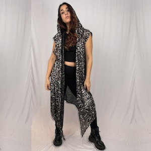 May include: A sleeveless, hooded duster coat with a black and silver animal print pattern. The long, open-front coat features a draped design and is paired with black leggings and boots. The coat's hood is up, and the fabric has a shimmering effect.